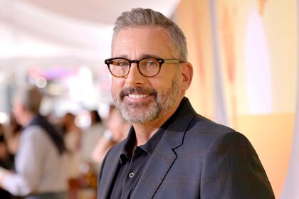 Steve Carell, wearing a black blazer and glasses, smiling and posing at an event.
