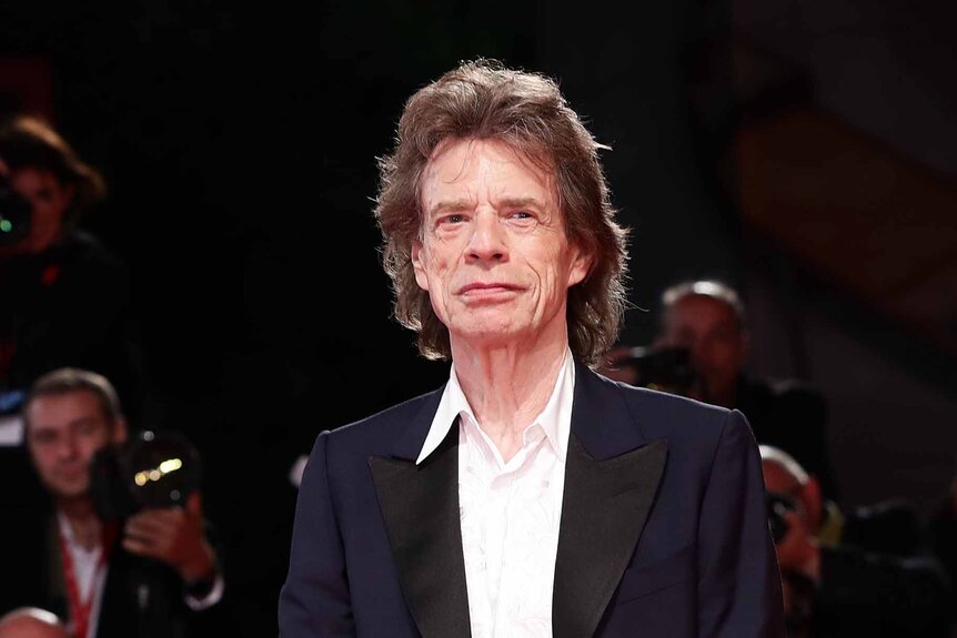 Mick Jagger wears a white shirt wiht a black jacket.