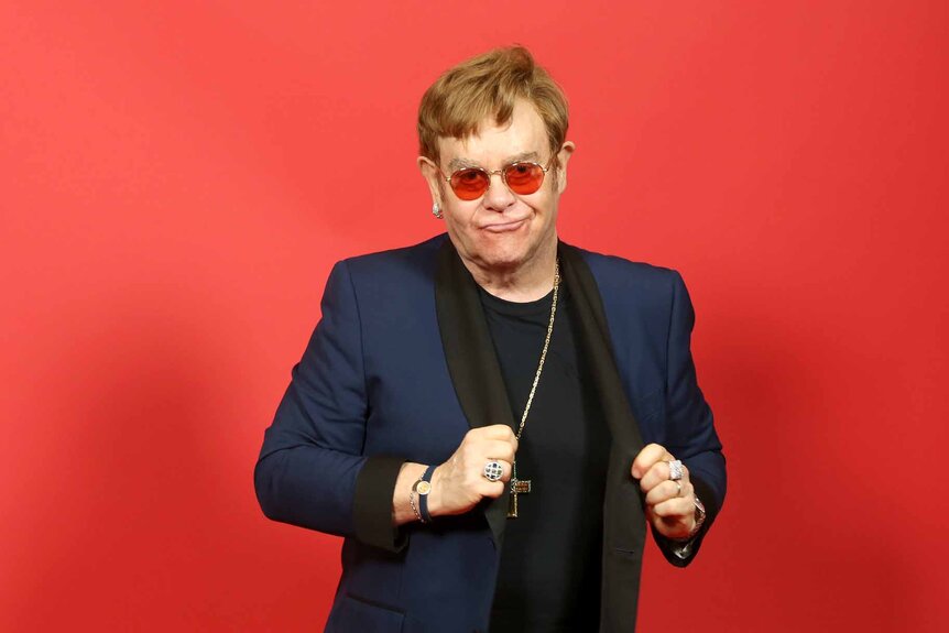 Elton John wears a blue jacket with red glasses in front of a red background.