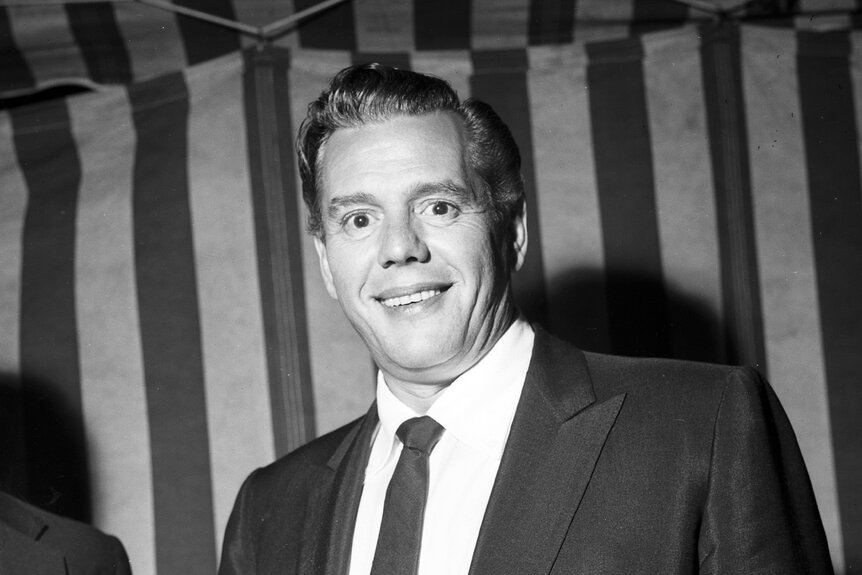 Desi Arnaz smiles in a suit in front of a striped background.