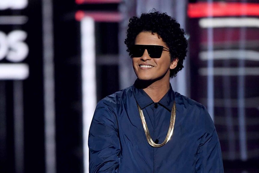 Bruno Mars smiles in sunglasses, a gold chain, and a blue shirt.