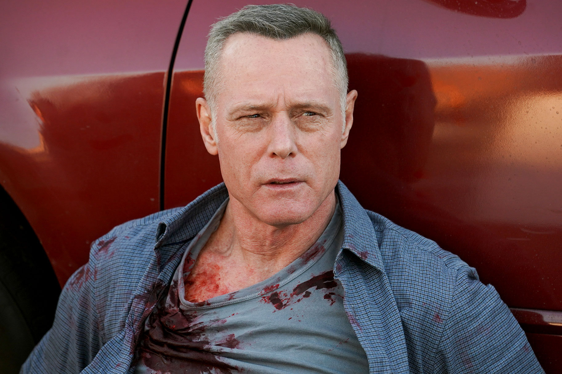 Jason Beghe sitting against a car with blood on his shirt