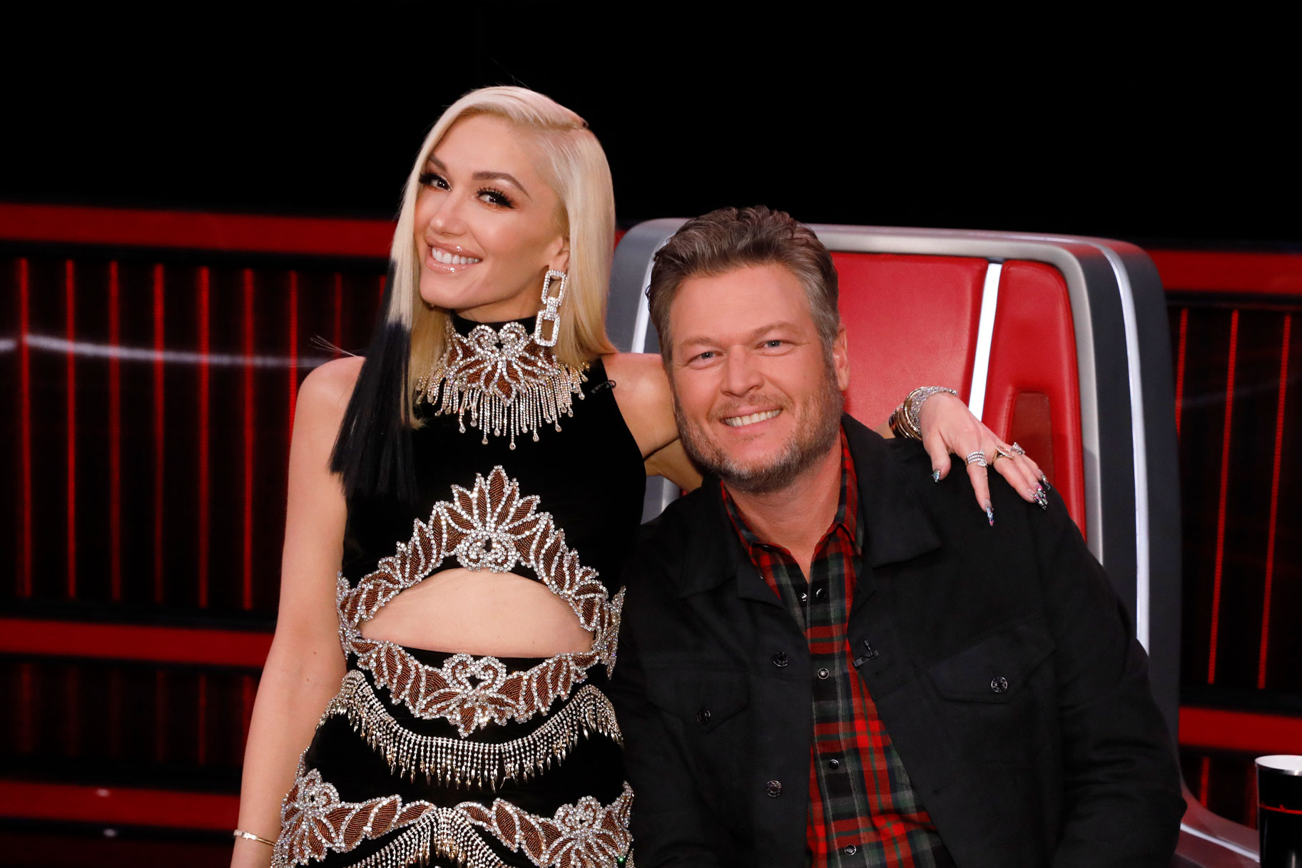 Gwen Stefani stands next to Blake Shelton who sits in his Voice chair. Both of them are smiling.