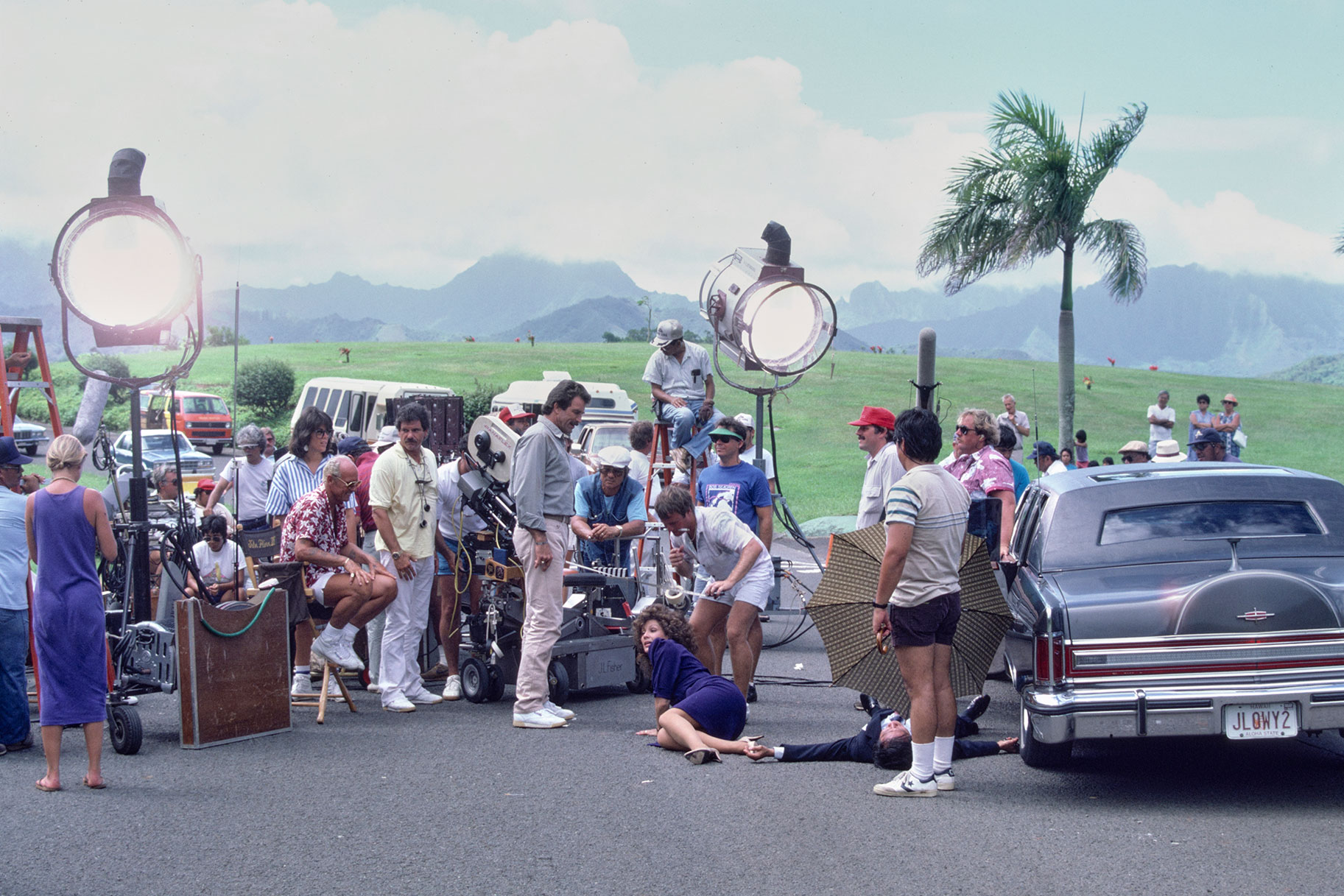 Tom Selleck (as Magnum) (in center wearing white pants) with Marta DuBois (as Michelle Hue) (in blue dress on the ground), in a production shot of the MAGNUM PI episode, "Little Girl Who."