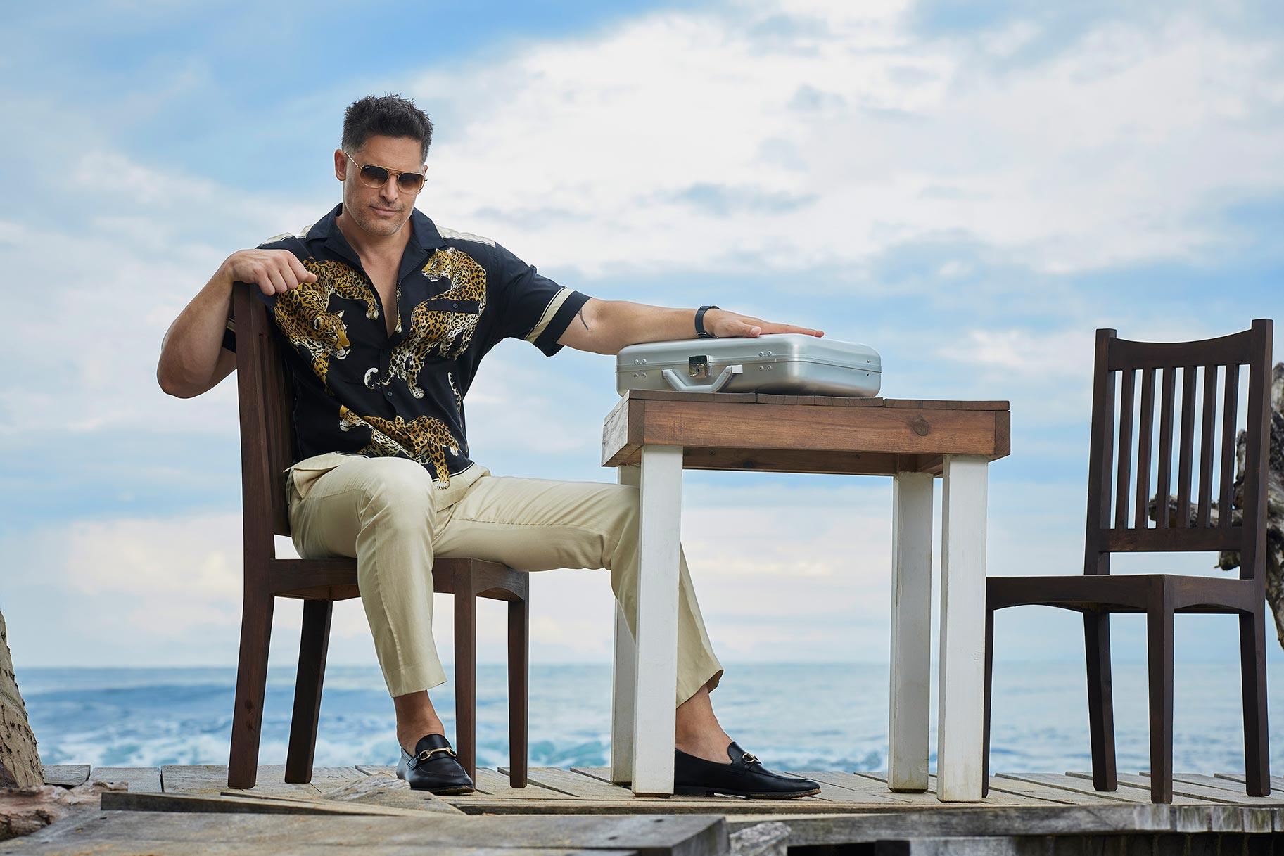 Joe Manganiello sits at a table on the beach with a silver briefcase