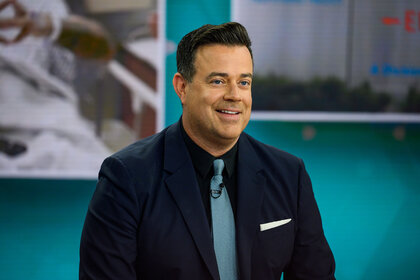 Carson Daly smiling while looking off camera. He's dressed in a dark blue suit.