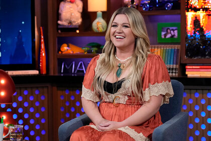Kelly Clarkson sits on a talk show couch, smiling.