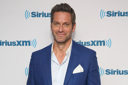 Peter Hermann smiles while standing in front of a SiriusXM step and repeat
