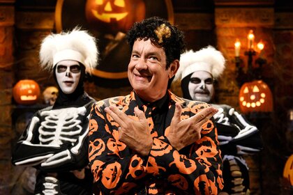 Tom Hanks dressed as character David S Pumpkins holding up two finger guns along with two skeletons beside him.
