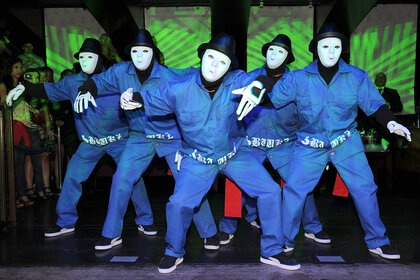 The dance group the Jabbawockeez pose in blue shirts and blue pants wearing hats and masks