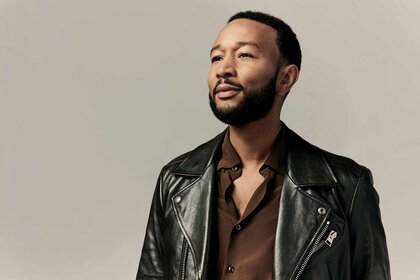 John Legend, wearing a brown top with a black leather jacket, looking off into the distance and posing for the camera.