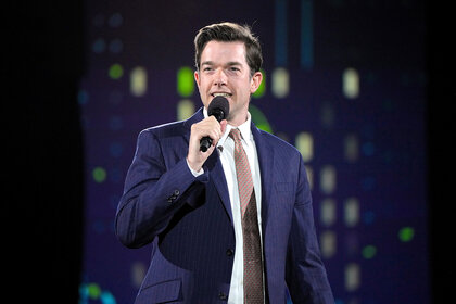 John Mulaney  performs on stage holding a microphone wearing a suit