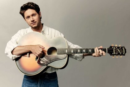 Niall Horan, wearing a white top, holds a guitar horizontally while posing for the camera.