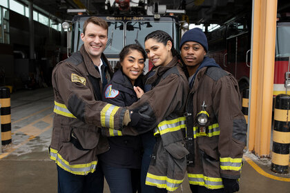 Matthew Casey, Gianna Mackey, Stella Kidd, and Darren Ritter hugging and posing for a photo in their firefighter jackets.