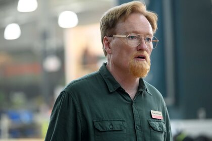 A close-up of Farley wearing a green collared shirt with glasses staring off into the distance.