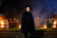 Alan Cumming stands in front of a mansion with flames on Season 2 of The Traitors