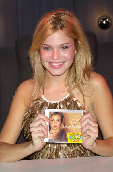 A young Mandy Moore sitting at a table and holding up her studio album.