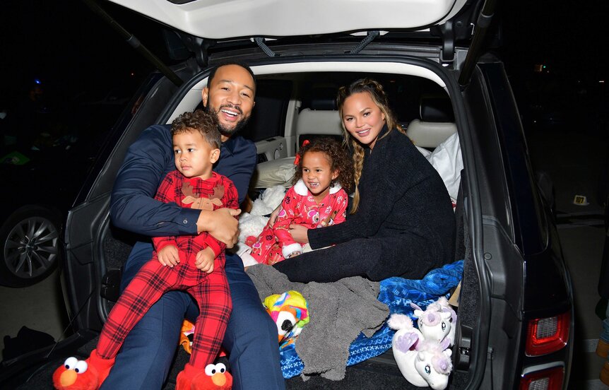 Miles Theodore Stephens, John Legend, Luna Simone Stephens, and Chrissy Teigen sitting in the trunk of a car posing for pictures.