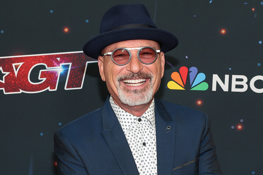 Howie Mandal on a red carpet for America's Got Talent smiling wearing glasses and a hat