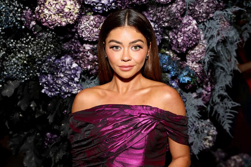 Sarah Hyland poses at an event wearing a purple top with a floral wall in the background.