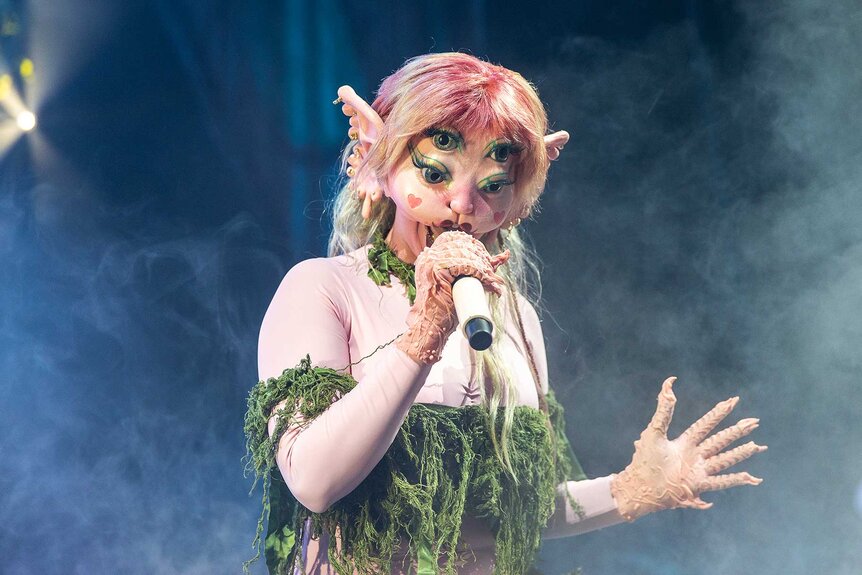 Melanie Martinez wearing a mask with multiple eyes and pink hair while performing.