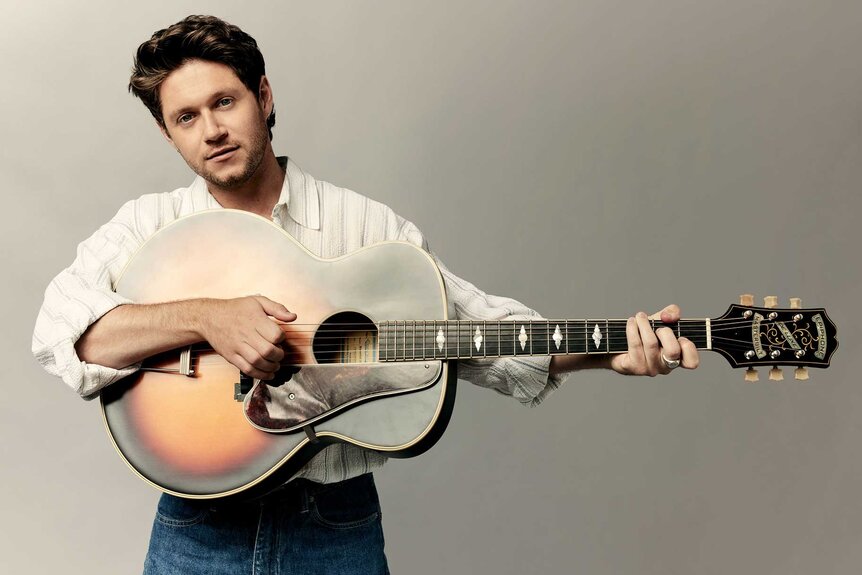 Niall Horan, wearing a white top, holds a guitar horizontally while posing for the camera.