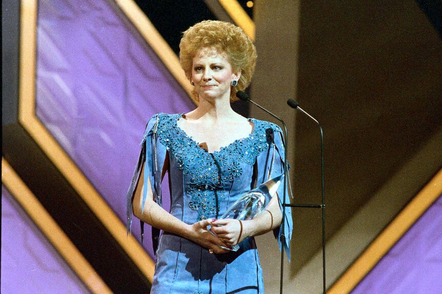 Reba McEntire standing on stage accepting an award wearing a blue dress.
