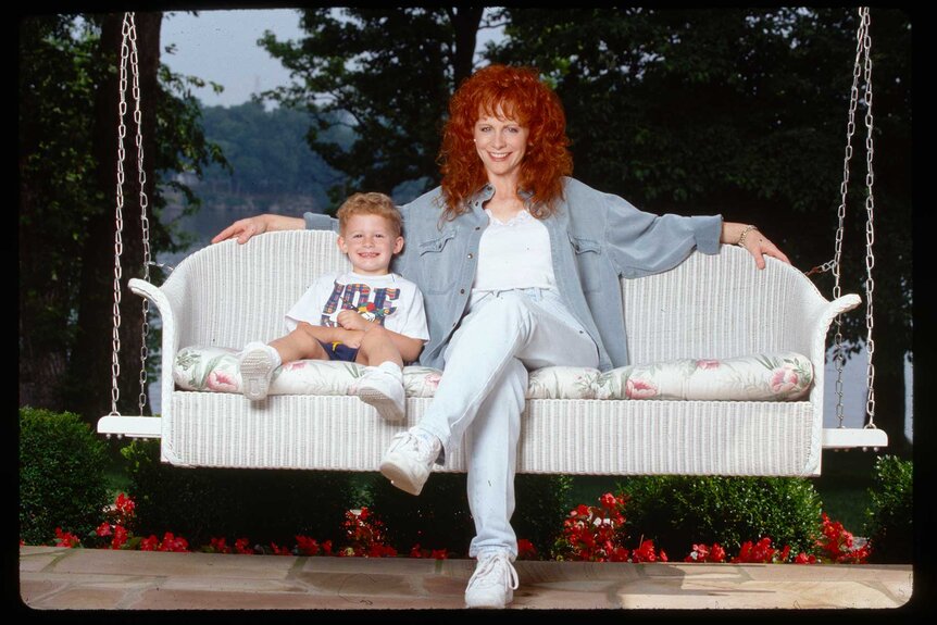 Shelby Blackstock as a baby posed next to his mom, Reba McEntire sitting on a white porch swing.