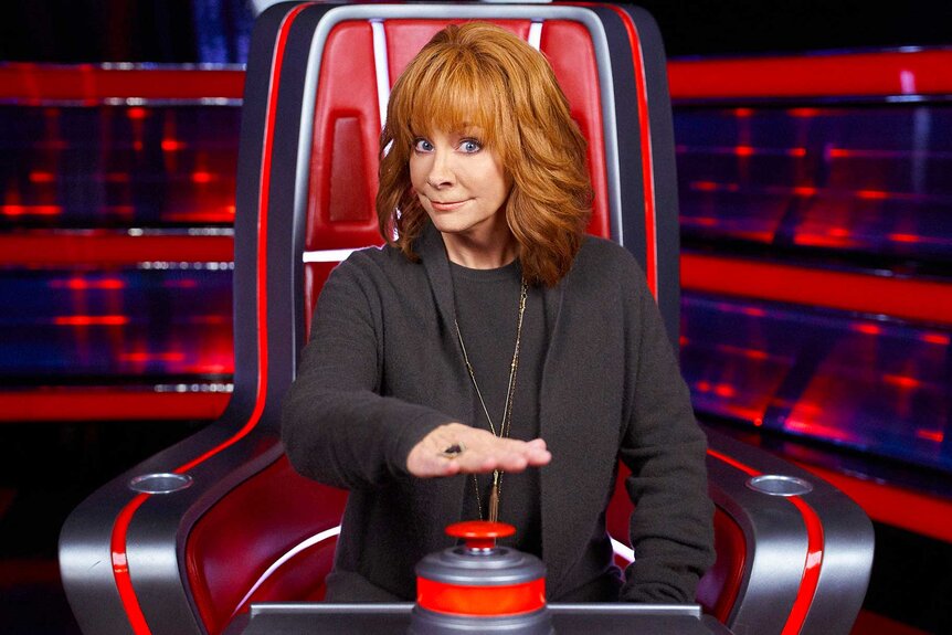 Reba McEntire in the middle of pushing a button while sitting in a judges chair for The Voice.