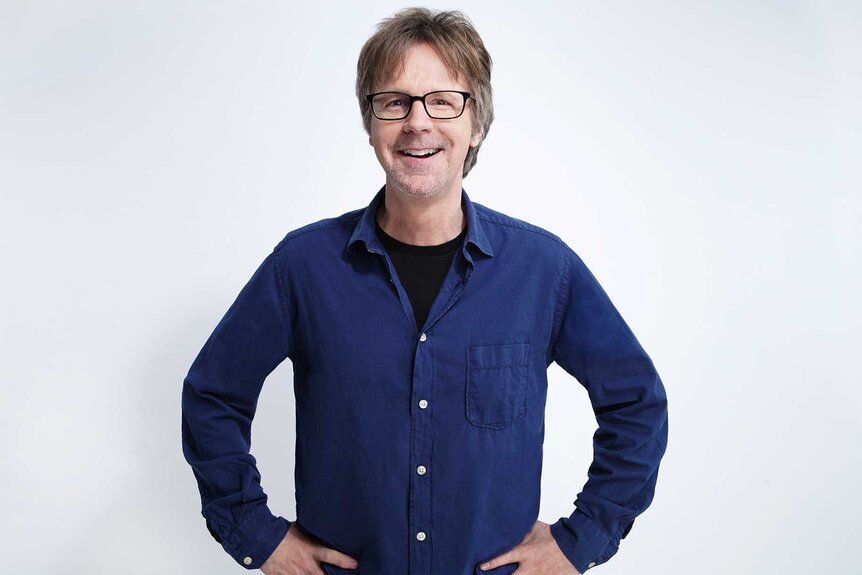 Dana Carvey posing with his hands on his hips wearing a blue shirt.