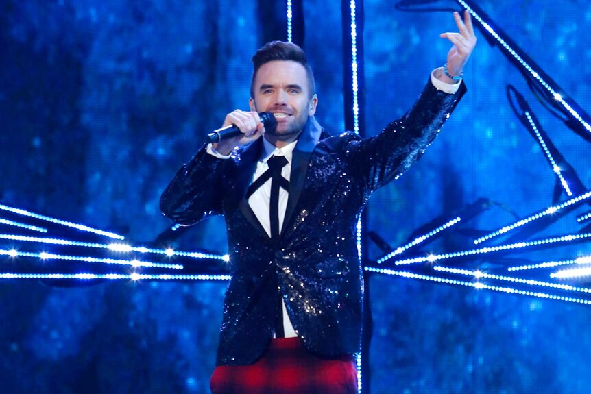 Brian Justin Crum, wearing a black blazer and plaid bottoms, on stage with a mic in his hand with his other arm raised.
