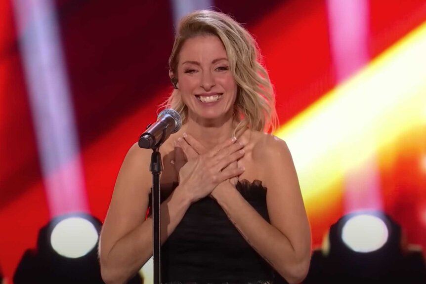 Genevieve Cote in front of a microphone with her hands on her chest while smiling.