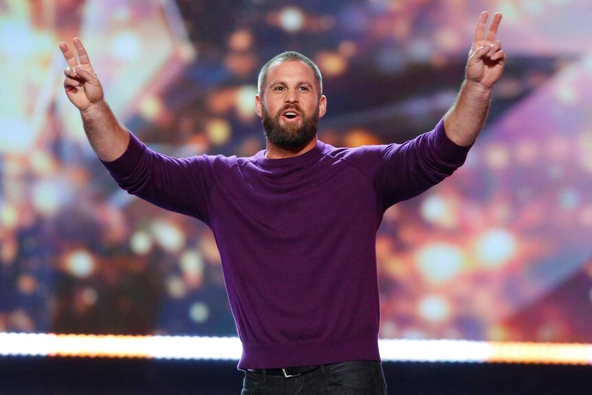 Jon Dorenbos, wearing a purple shirt, on stage holding up two peace signs.