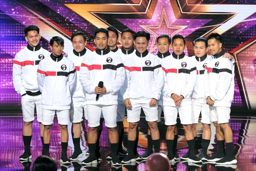 Junior New System, wearing white uniforms and black shoes, grouped together on stage.