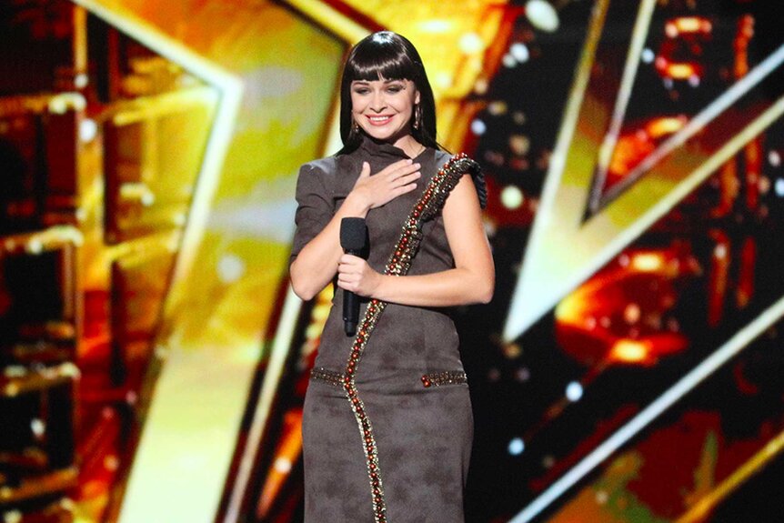 Kseniya Simonova, wearing a gray dress, stands on stage holding a microphone in one hand while putting her other hand on her heart.