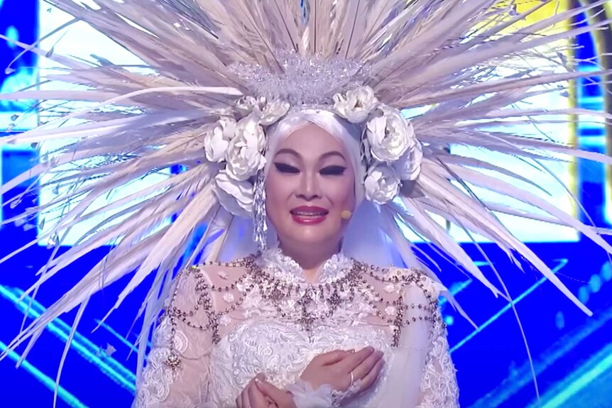 A close up of Maria Seiren, wearing a large, all white, feather headpiece on stage.