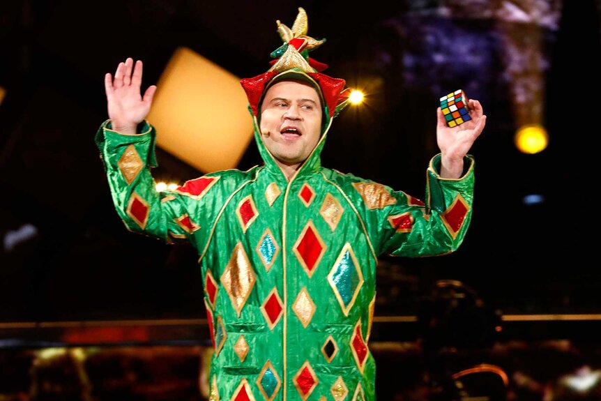 Piff The Magic Dragon wearing a green, red, and gold costume while holding a rubik's cube.