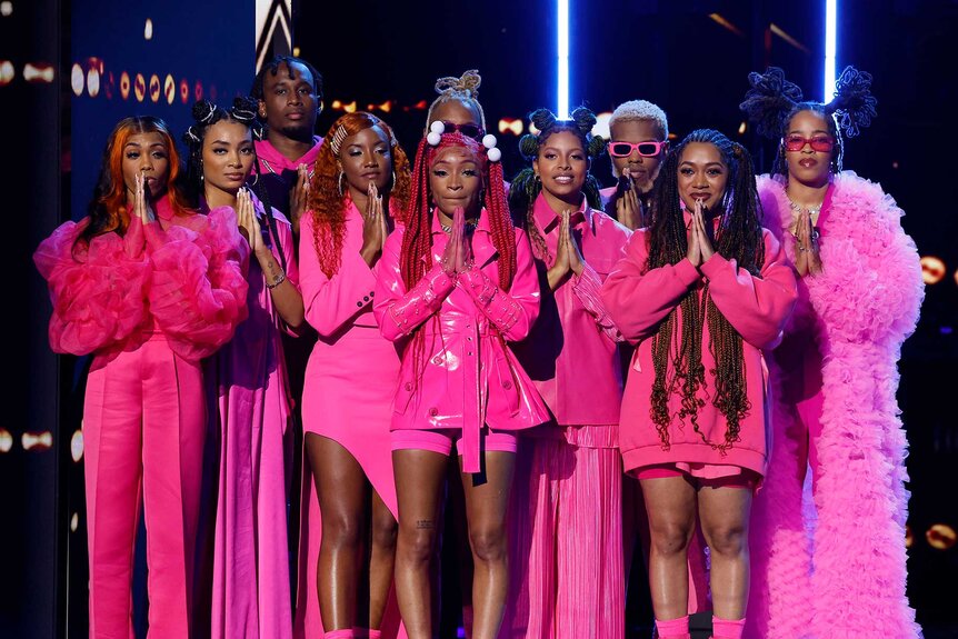 Sainted, wearing all pink outfits, standing together on stage with each group member putting their hands together in a prayer gesture.