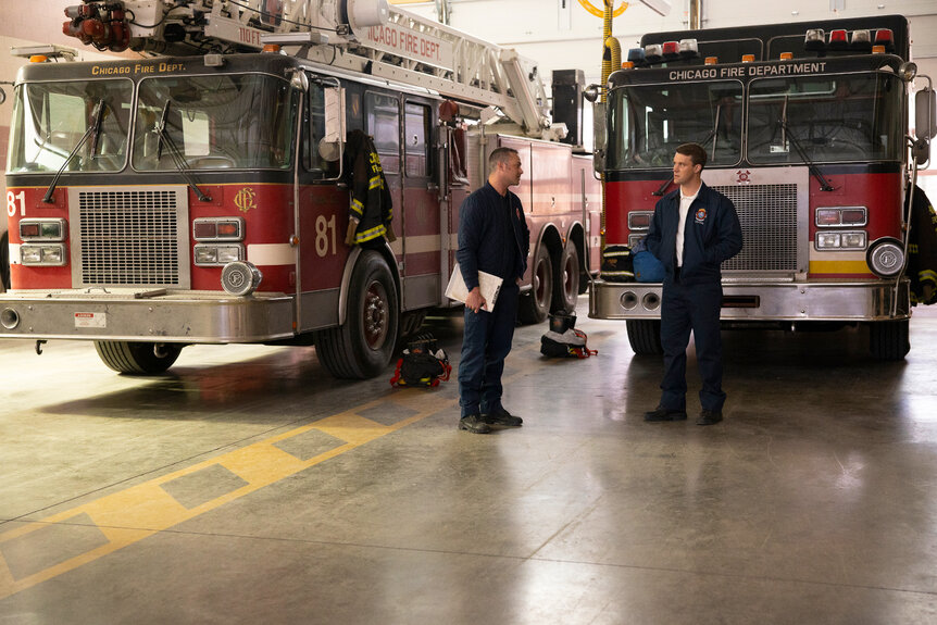 Kelly Severide standing with Matthew Casey in front of two firetrucks.