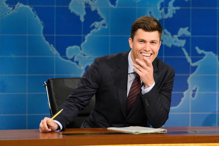 Colin Jost wears a gray suit with his hand touching his chin at his desk during the Weekend Update segment.