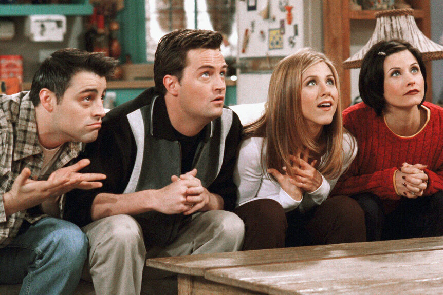 The cast of friends sit on a couch looking excited