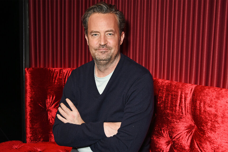 Matthew Perry sits on a red couch with his arms crossed