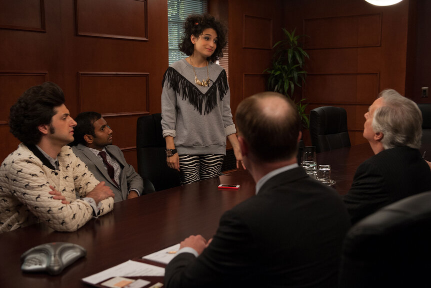 Jenny Slate as Mona-Lisa Saperstein stands while speaking to a table of men