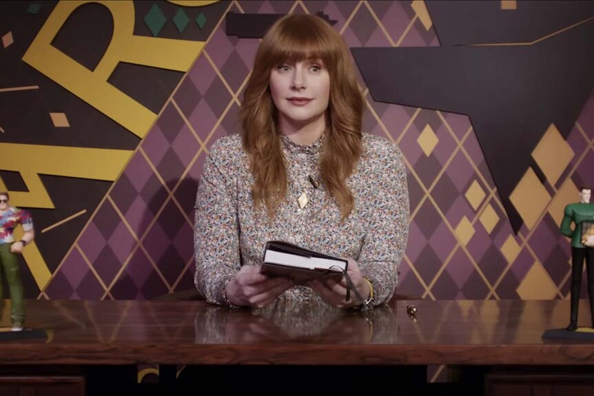 Bryce Dallas Howard holds a book at a table in front of a background of purple and yellow argyle in Argylle (2024).