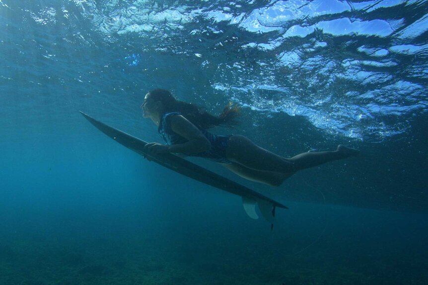 Malia Manuel underwater with a surfboard in hand.
