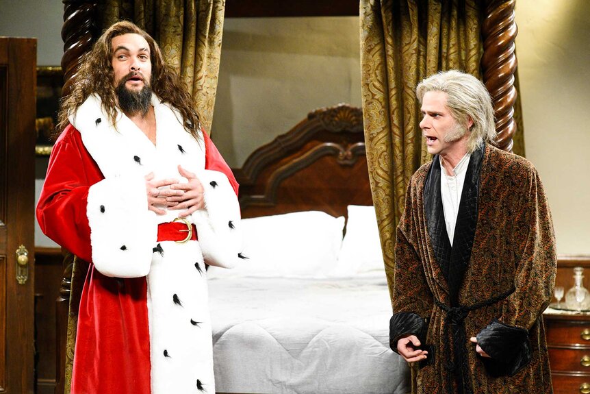Jason Momoa dressed in a Santa coat while standing next to Mikey Day dressed as Ebenezer Scrooge during Saturday Night Live.