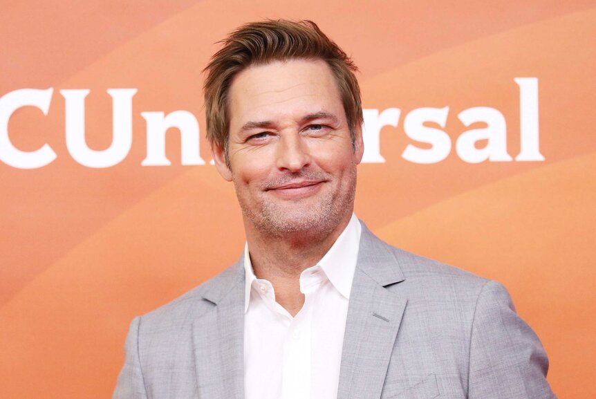 A close up of Josh Holloway wearing a grey suit and posing in front of an orange backdrop.