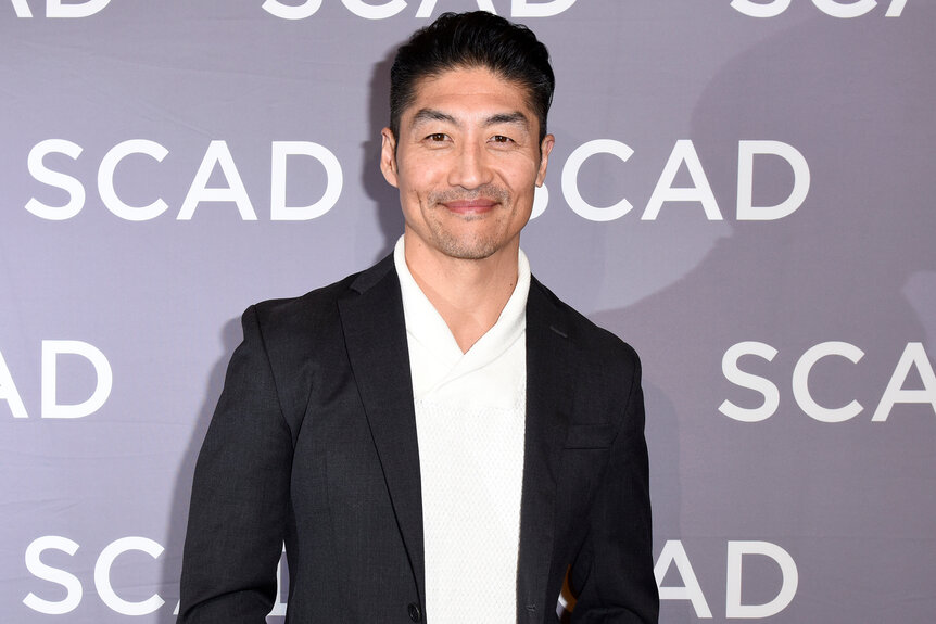 Brian Tee smiles in a black suit with white shirt on the red carpet at a panel for The Windy City Trifecta: Dick Wolf's 'Chicago'