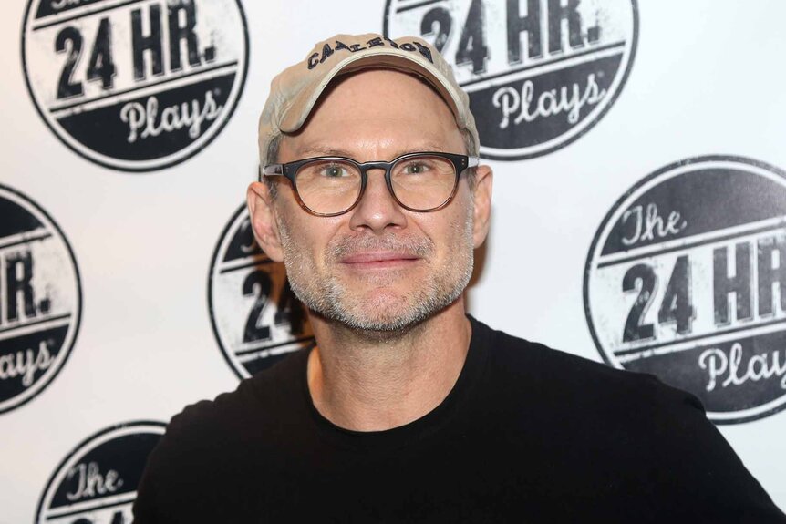 Christian Slater smiles in glasses and a cap.