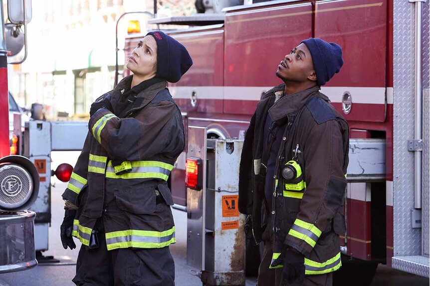 Stella Kidd and Darren Ritter look up while wearing firefighter uniforms near a fire truck in Chicago Fire Episode 1202.